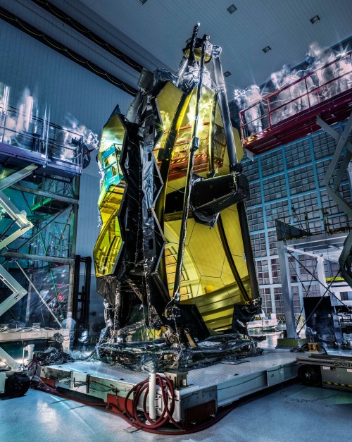 The image shows the James Webb Space Telescope in a clean room, illuminated with colorful lights and intricate machinery.