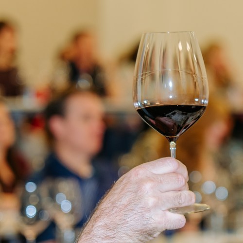 A hand holding a glass of red wine, with blurred people in the background at a gathering or event.