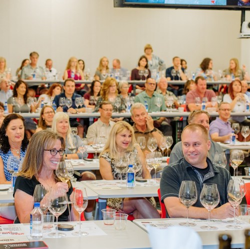A large group of people seated at tables, enjoying a wine tasting event with glasses and bottles in front of them.
