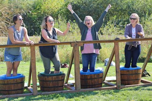 Four women stand in wooden barrels, smiling and celebrating outdoors on a sunny day. Greenery surrounds them.