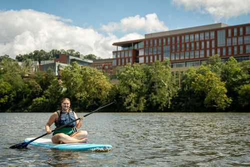 A person sits on a paddleboard in a river, smiling, with trees and a modern building in the background.