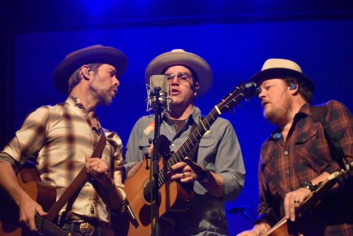 Three musicians in hats perform together on stage, singing into a microphone with guitars in hand, under blue lighting.