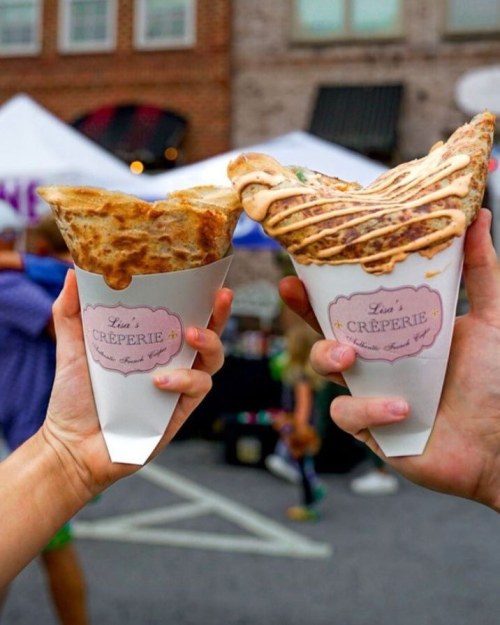 Two hands hold delicious crepes in paper cones from Lisa's Crêperie, with a festive background.