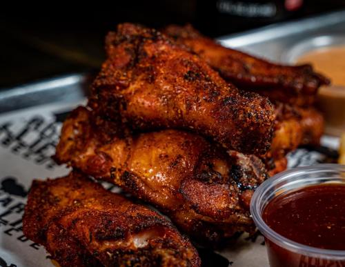 A close-up of crispy, seasoned chicken wings stacked on a tray, accompanied by a small cup of barbecue sauce.