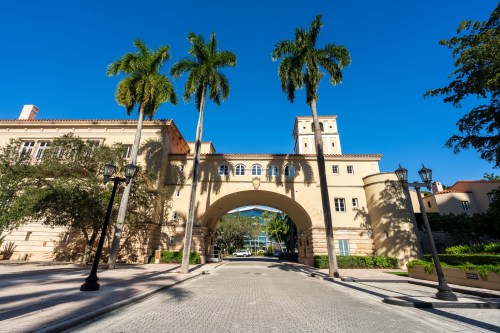 A sunny street view featuring palm trees and an archway between two buildings. Clear blue sky above.