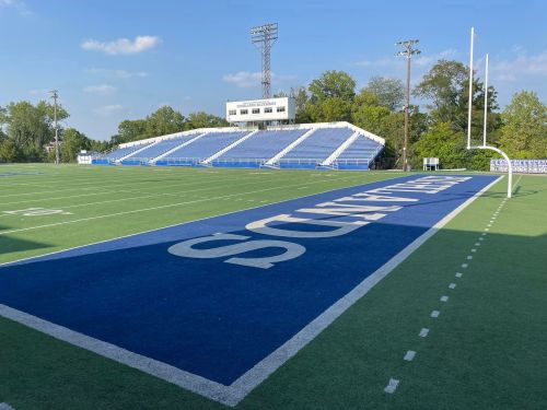 A football field with blue turf, marked yard lines, and a grandstand in the background under a clear sky.