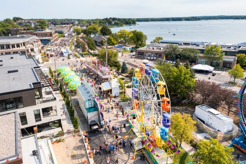 Aerial view of a lively festival by a lake, featuring a Ferris wheel, colorful tents, and crowds enjoying the event.