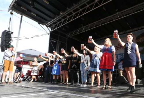 A group of people in traditional attire raise beer steins on stage during a festival celebration.
