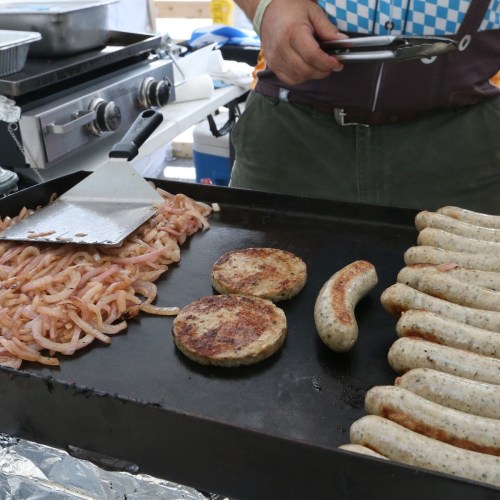 A person cooking sausages and patties on a griddle, with onions sautéing nearby.