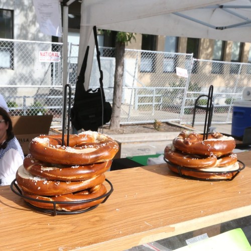 A display of large, soft pretzels stacked in a wire basket on a wooden table at an outdoor market.