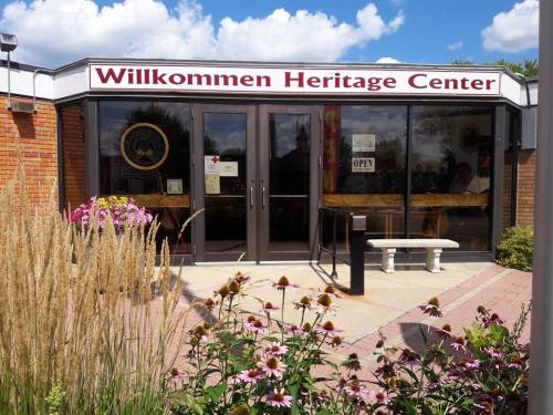 Entrance of the Willkommen Heritage Center, featuring flowers and a sign that says "Open" under a blue sky.