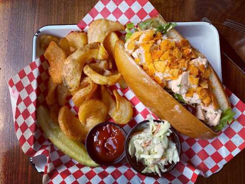 A sandwich filled with shredded chicken and topped with crushed chips, served with curly fries, coleslaw, and pickles.