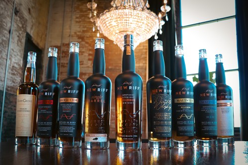 A lineup of New Riff whiskey bottles displayed elegantly under a chandelier in a rustic setting.