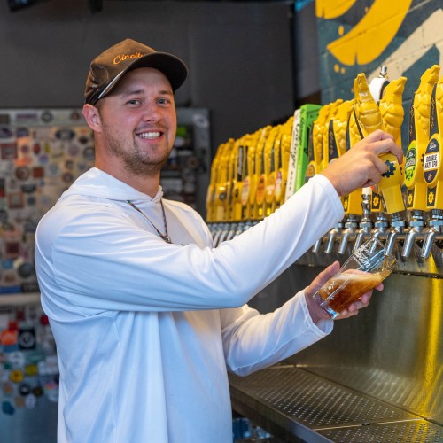 A smiling man in a white shirt pours a drink from a tap at a bar with colorful beer handles.