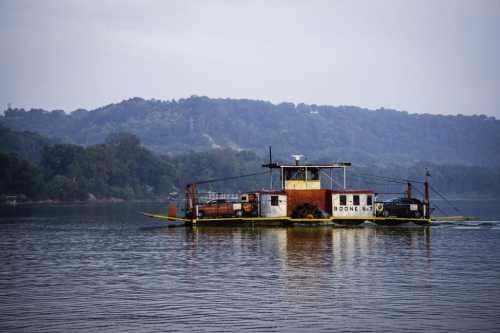 A ferry boat with cars on board navigates a calm river, surrounded by green hills under a cloudy sky.