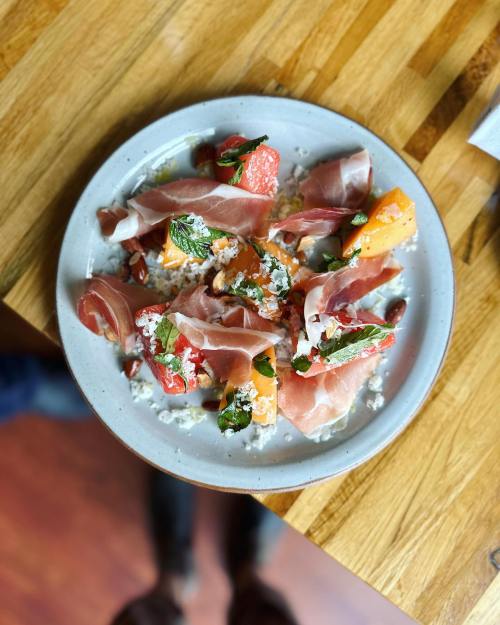 A plate of colorful salad with melon, tomatoes, mint, and slices of prosciutto on a wooden table.