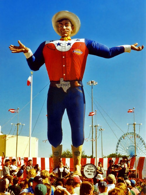A large cowboy statue in a red, white, and blue outfit stands over a crowd at a fair, with a ferris wheel in the background.