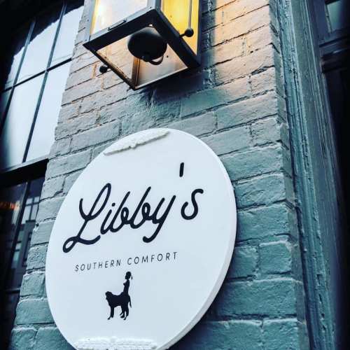 Sign for "Libby's Southern Comfort" with a dog silhouette, mounted on a textured wall beside a lantern.