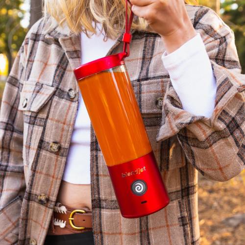 A person holds a red portable blender with an orange smoothie inside, wearing a plaid jacket outdoors.