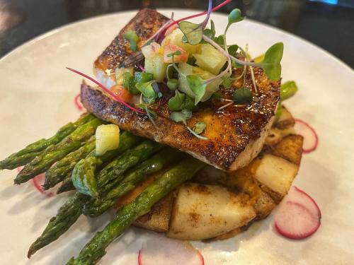 A beautifully plated dish featuring grilled fish atop asparagus and potatoes, garnished with fresh salsa and microgreens.