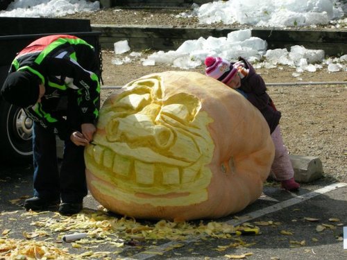 A child watches as an adult carves a large, smiling pumpkin in a snowy outdoor setting. Pumpkin guts are scattered around.