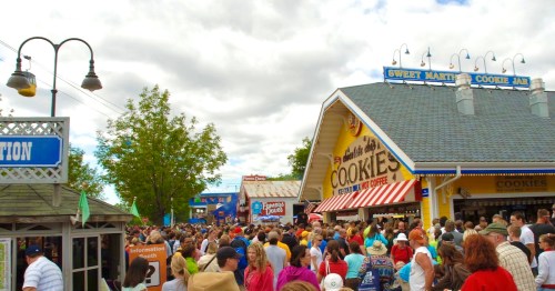 A crowded outdoor fair with people walking past cookie stands and colorful signage under a cloudy sky.