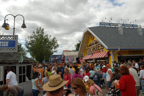 A bustling fair scene with a large crowd outside a cookie shop under cloudy skies.