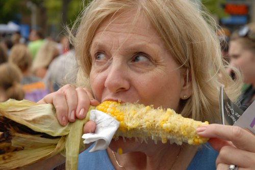 A woman bites into a cob of corn, surrounded by a crowd at an outdoor event.