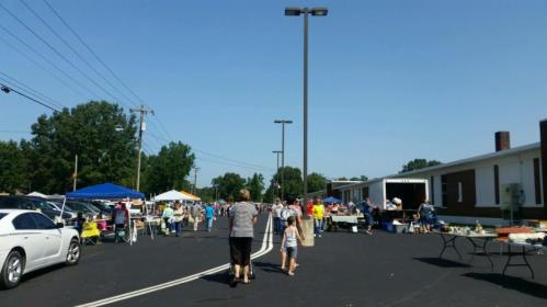 A bustling outdoor market with people browsing stalls under a clear blue sky. Cars and tents are visible along the pathway.