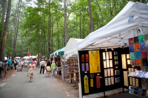 A vibrant outdoor market in a forest, featuring art displays and visitors exploring various booths among tall trees.