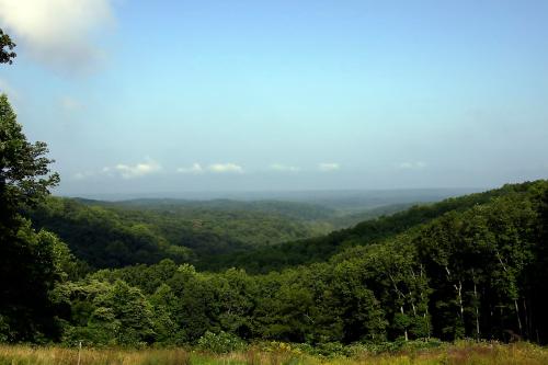 A panoramic view of lush green hills under a clear blue sky, with distant mountains fading into the horizon.