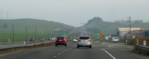 A highway scene with cars traveling on a multi-lane road, surrounded by green hills and distant power lines.