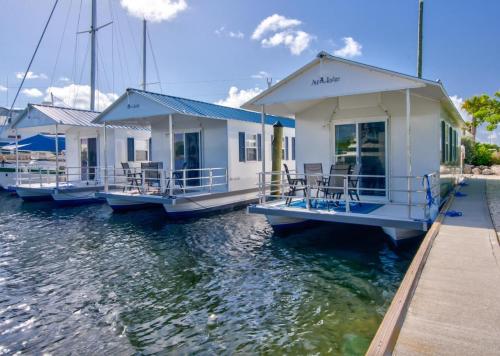 Three white houseboats with porches docked on calm water, surrounded by blue skies and a few sailboats in the background.