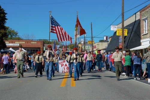 A parade with participants holding flags, marching down a street lined with spectators and shops.