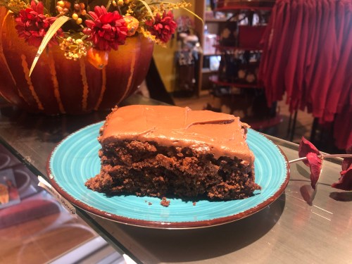 A slice of chocolate frosted cake on a blue plate, with a decorative pumpkin and flowers in the background.