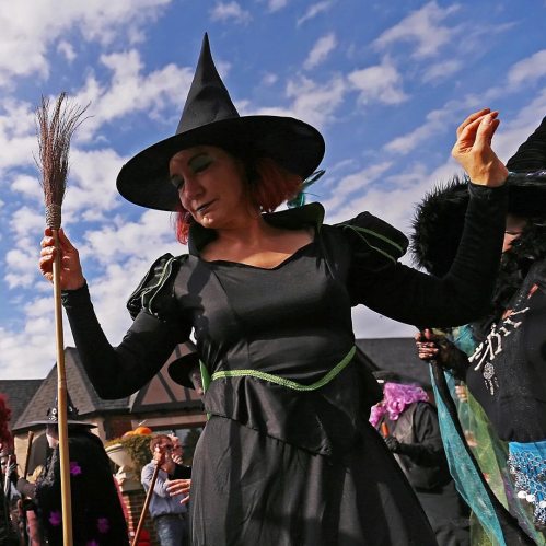 A woman in a black witch costume and hat dances with a broom, surrounded by a festive crowd under a blue sky.
