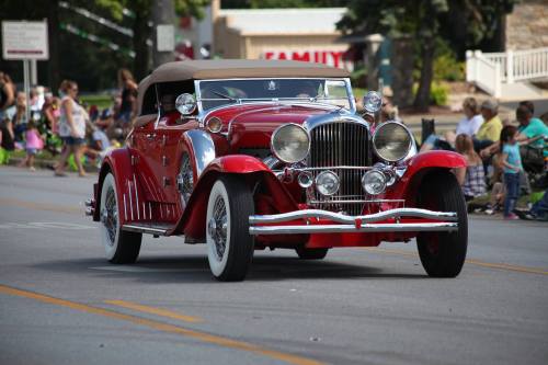 A vintage red convertible car with a tan top drives down a street, surrounded by spectators at a parade.