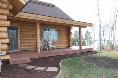 A log cabin with a porch, featuring two chairs, surrounded by grass and trees near a lake.