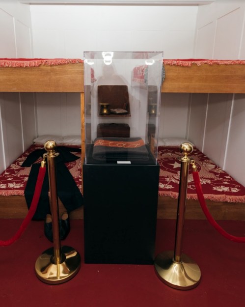 Display case with a red item inside, surrounded by velvet ropes, in a room with two wooden bunks.
