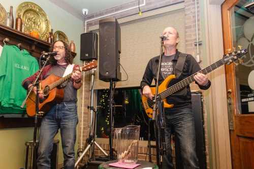 Two musicians perform live in a cozy venue, one on guitar and the other on bass, with a microphone and speakers nearby.