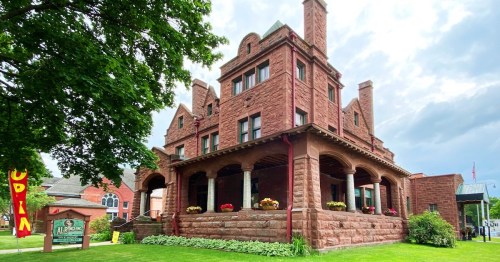 Historic red stone building with a porch, surrounded by greenery and flowers, under a cloudy sky.