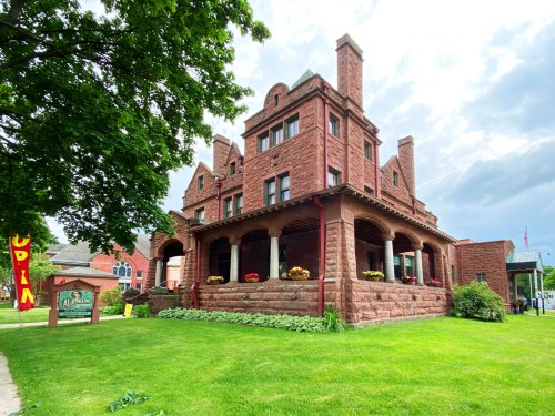 Historic red stone mansion with a porch, surrounded by green grass and trees, under a cloudy sky.