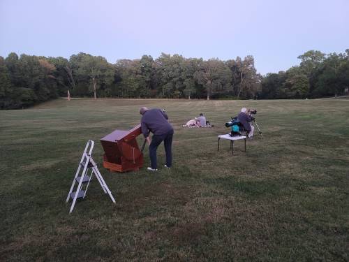 Two people set up equipment on a grassy field at dusk, with trees in the background and a ladder nearby.