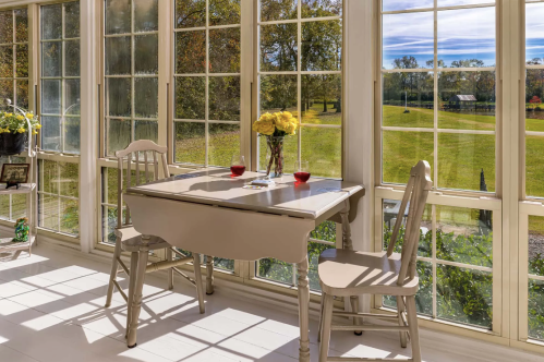 A bright sunroom with a small table, two chairs, and large windows overlooking a green landscape. Yellow flowers on the table.