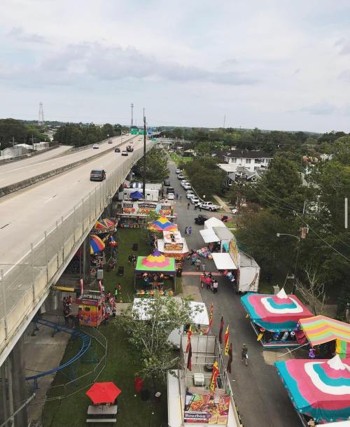 Aerial view of a fair with colorful tents and booths alongside a highway, surrounded by trees and buildings.