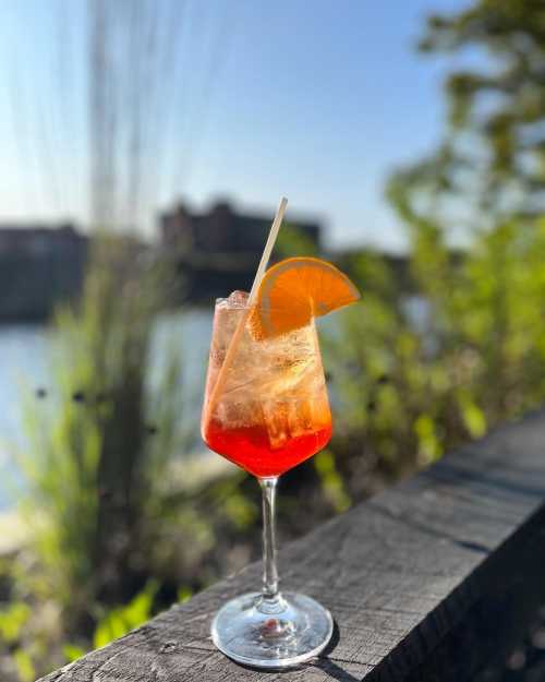 A refreshing cocktail with orange slice, served in a glass, against a scenic riverside background.