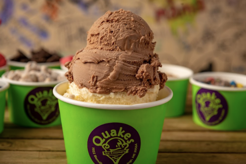 A close-up of a chocolate ice cream scoop atop vanilla in a green cup from Quaker Ice Creamery, with colorful toppings in the background.