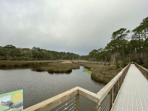 A wooden boardwalk stretches over a calm marsh, surrounded by trees and cloudy skies.