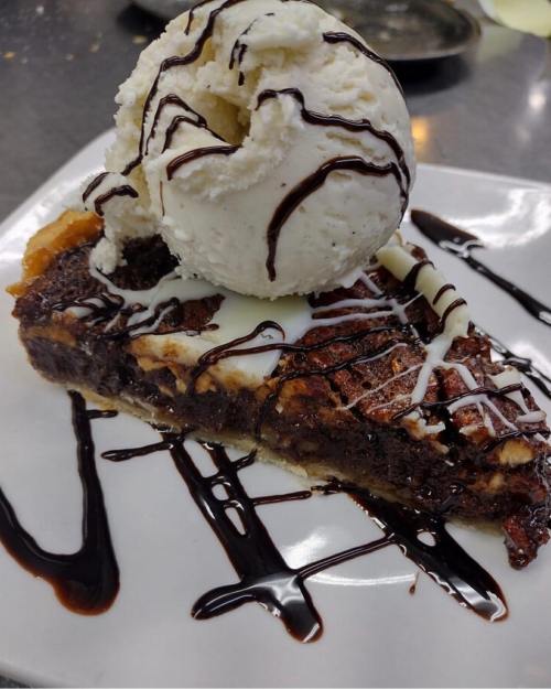 A slice of chocolate brownie pie topped with a scoop of vanilla ice cream and drizzled with chocolate sauce.