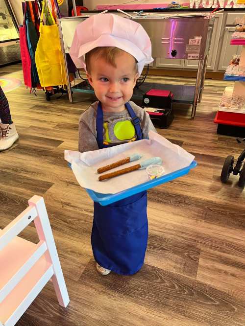 A young child in a chef's hat and apron holds a tray with pretzels and dipping sauce, smiling happily.